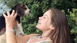Happy teenage girl holding up a brown and white Beagle puppy outside.